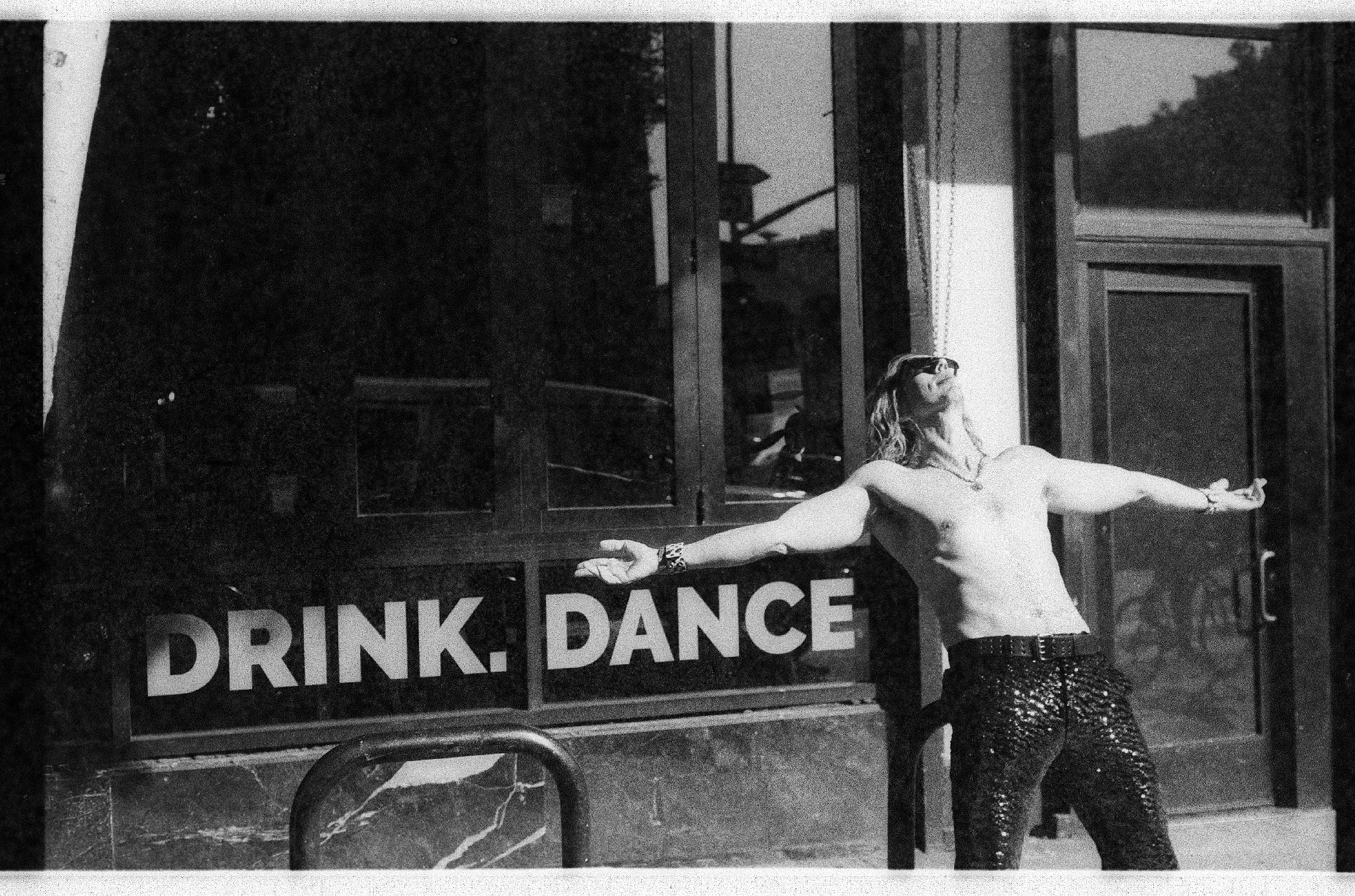 Indiana Bradley outside a storefront reading DRINK. DANCE, arm outstretched, in black and white
